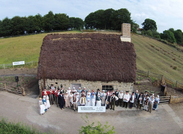 aerial shot of stone cottage with heather thatch with lots of people in 18th century dress