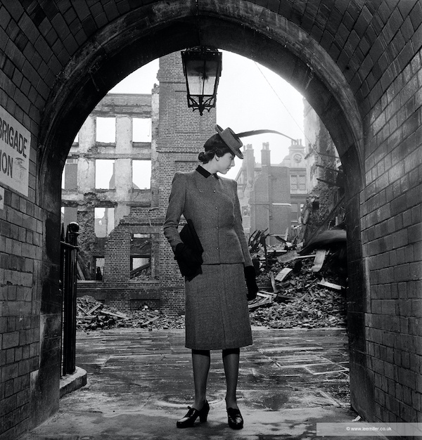 A model stands in a brick archway facing forwards but looking down to her left. Behind her there is bomb damage buildings broken and bricks on the ground. She wears a smart suit and hat and wears gloves with a handbag. The black and white photograph is by Lee Miller.