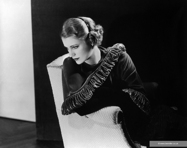 A black and white photograph of Lee Miller. A self portrait, a woman sits in a chair leaning over the side looking down to the left she wears a black outfit and long black gloves.