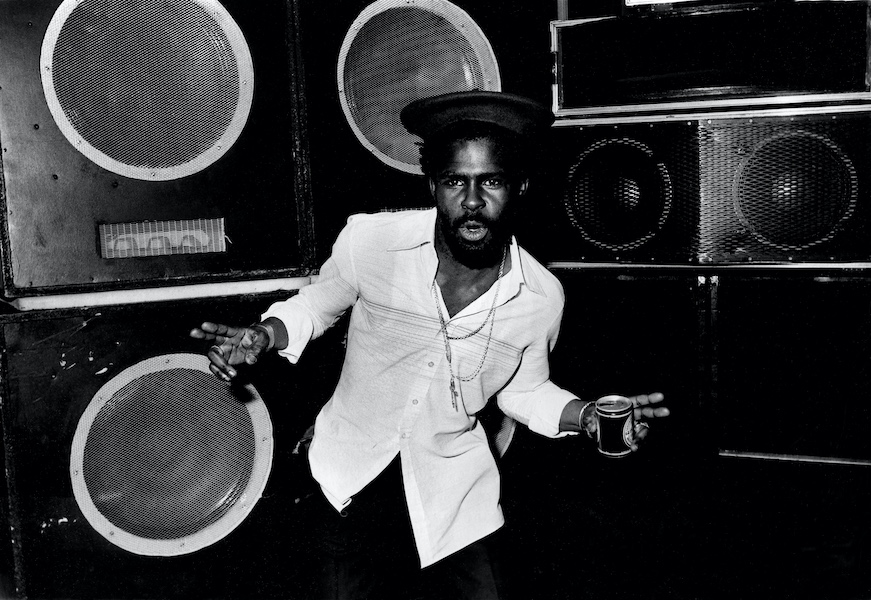 A black and white photograph of a black man who is dancing in front of a sound system of large speakers. He has on a white shirt, black trousers and black hat and chains round his neck. In his left hand is a small can of guinness. He is facing the camera.