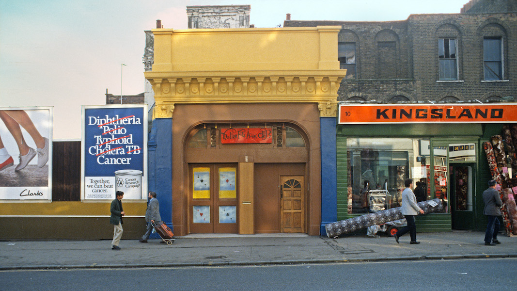 A colour photograph of a London street, facing the buildings, in the middle is a yellow and brown entrance to the Four Aces club, on the left is advertising hoarding and on the right a shop called Kingsland. People walk past in front of the buildings. 