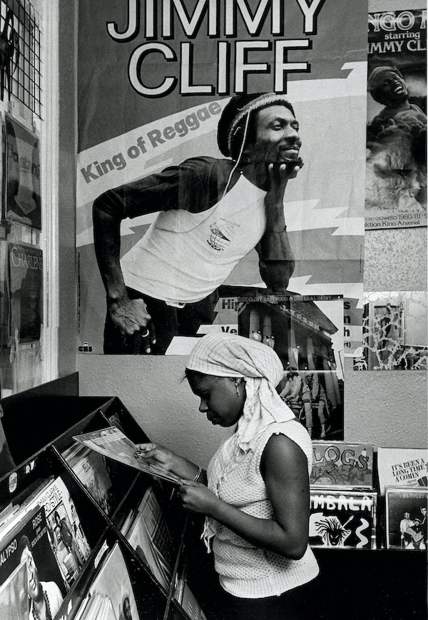 A black and white photograph of a black girl in a record shop, she stands side on looking down to her left as she selects records. Above her on the wall is a poster of the 'King of Reggae' Jimmy Cliff.