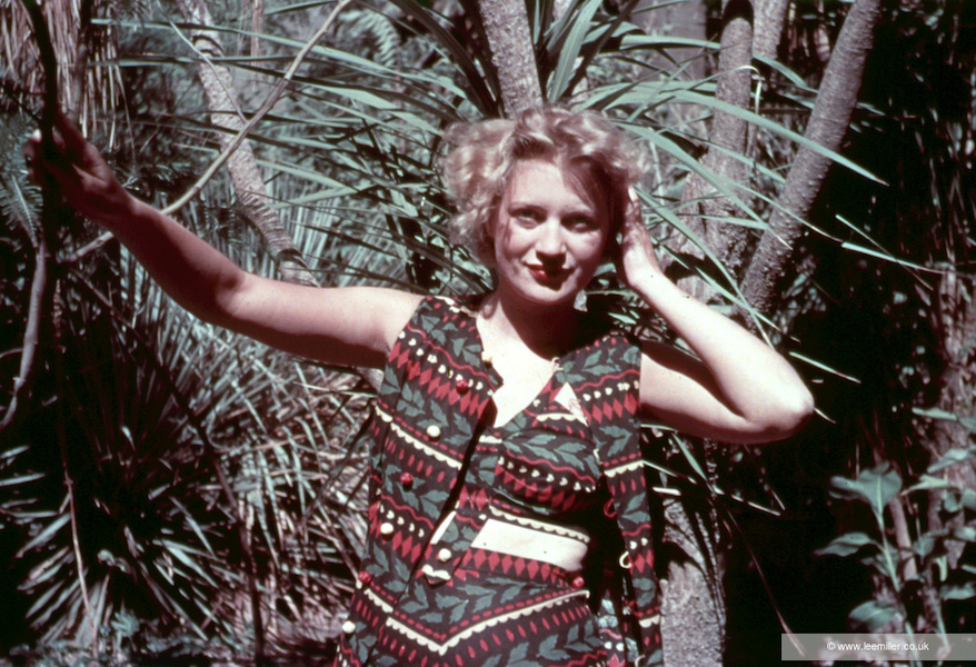 A colour photograph of Lee Miller, a blonde woman stands outside with green trees and vegetation looking directly at the camera her right arm raised resting on a tree. She wears a bra top under a short waistcoat and skirt. It is in a vibrant green and red pattern.