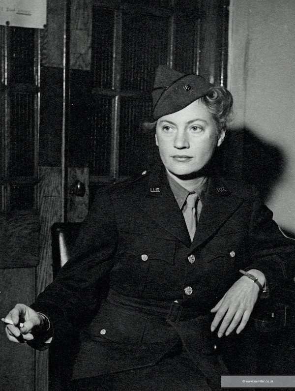 Lee Miller dressed in military uniform. A woman faces the camera looking off to the left she leans on her left elbow with a cigarette in her right hand. She wears a military uniform and hat. Her hair is tucked under her hat. It is a black and white photograph. 