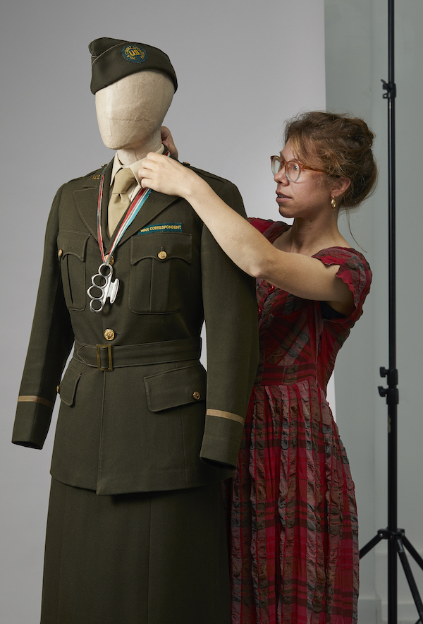 An installation photograph from the exhibition, a woman in a red dress stands behind a mannequin adjusting the neckline of the outfit on the mannequin. The outfit is a green military uniform with dark green skirt and jacket and a khaki shirt and tie. There is also a dark green hat.