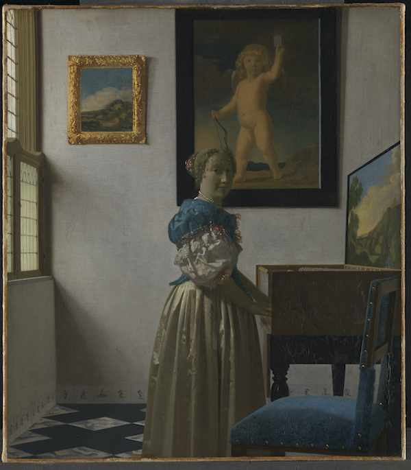 The painting by Vermeer shows a young woman standing in the middle of a room, she faces a virginal (keyboard) which her hands are resting on. Her head is turned to look at the viewer. The floor has black and white tiles, light comes in from a window on the left. She wears a light gold long skirt, white blouse with a blue shawl on her shoulders. Her dark brown hair is worn up in a bun. She also wears a pearl necklace. Behind her on the white wall are two paintings, in the foreground there is a high backed chair with a blue seat. 