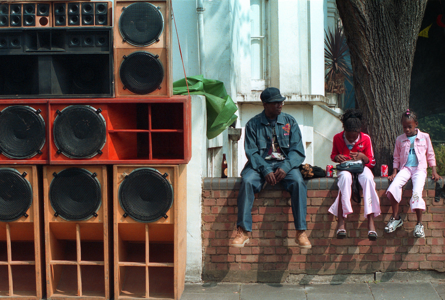 A colour photograph of the Notting Hill Carnival, the left hand of the image is full of a large stack of speakers, on the right of the image is a beige brick wall in front of a house on the wall sits a black man dressed in denim with a denim hat, next to him on the wall sits two young black girls dressed in pink and red. They have cans of coke next to them.