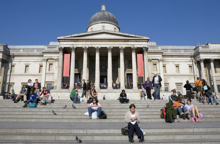 A photograph of the  National Gallery during the day. There is blue sky above the roof of the gallery. The steps are shown leading up to the entrance portico. Lots of people sit on the steps on their own and in groups. There are pigeons dotted about on the steps.  