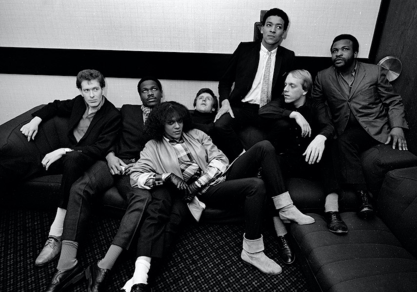 A black and white photograph of the band - The Selector. Seven band members including singer Pauline Black sit across a sofa squeezed togther. They all look in different directions, only Pauline looks at the camera.  