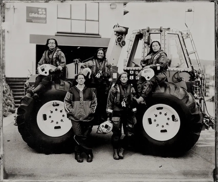 A black and white photo of five women by an RLNI tractor. Two women stand in front and three women sit on top of the tractor staring at the camera. They are wearing their waterproof RLNI clothes and three carry RLNI white helmets.