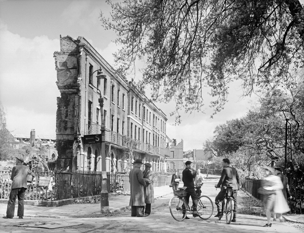 40s street with people chatting and one holding bicycle in front of destroyed building - only the frontage still stands