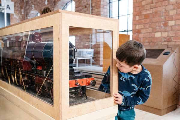 small boy in dinosaur top looks at large model steam train in glass case