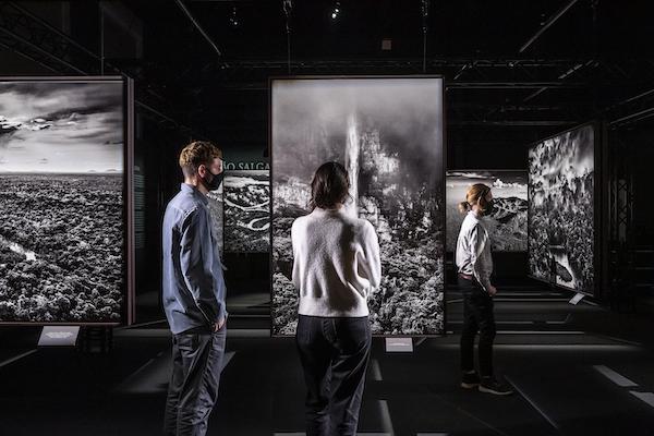 three people stand and look at large black and white photographs of the Amazon in darkened gallery