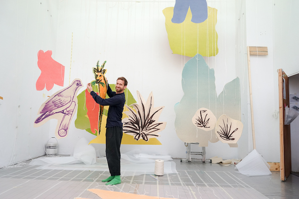 the artist stands surrounded by illustrations of a bird and antelope in his studio