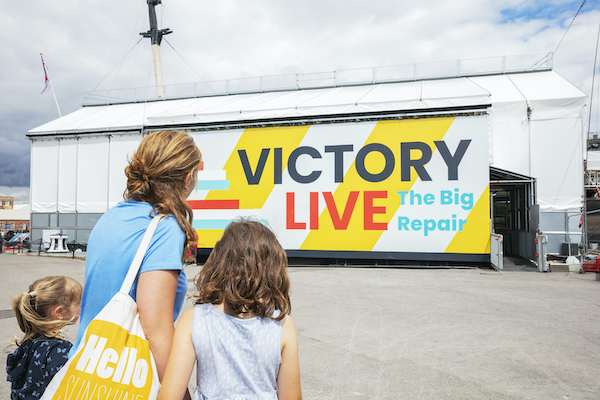 shed with victory live written on it with ship within and two visitors looking on
