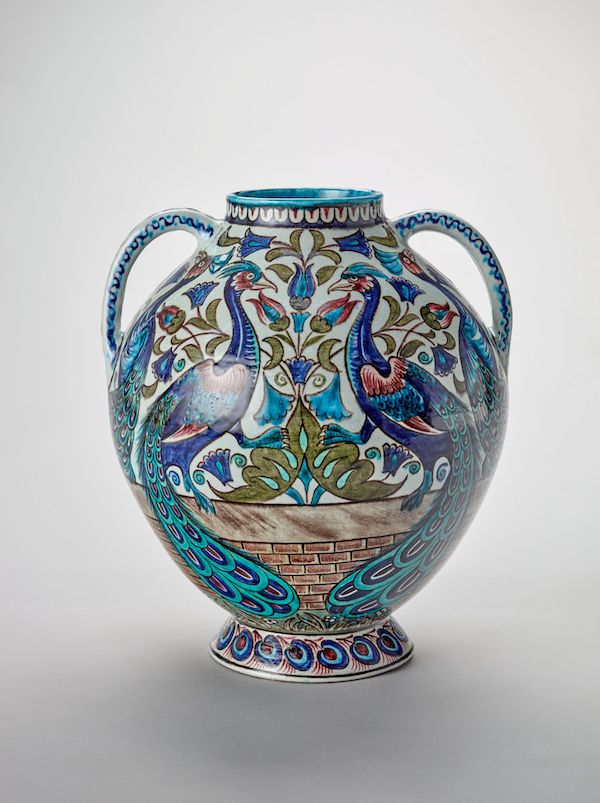 A photograph of an earthenware pottery vase that has two handles on either side. It has an intricate pattern in blues and greens of two peacocks sitting on a wall facing each other. The rim and handles are decorated with repeating patterns.  