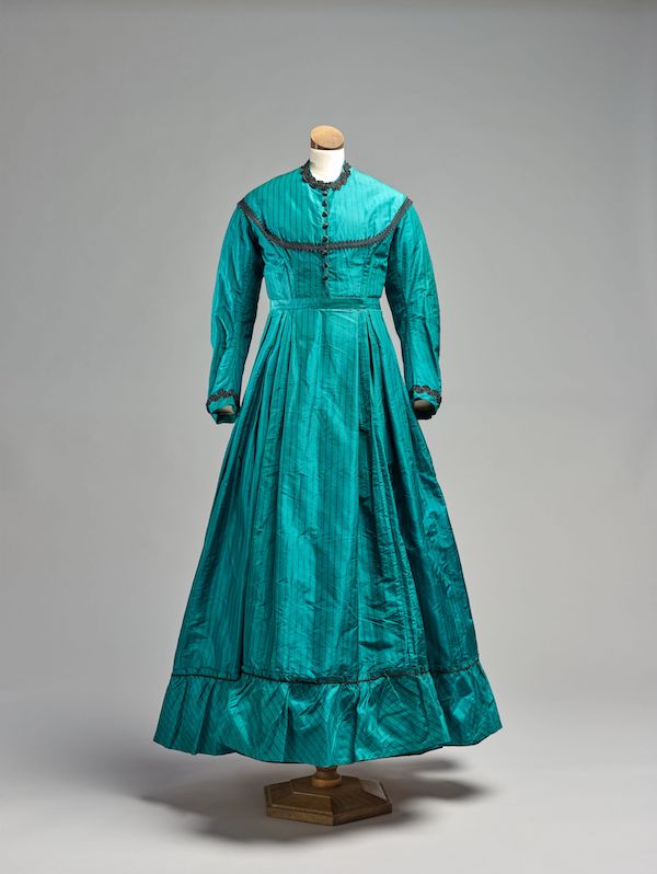 A mannequin displays a Victorian dress it is in a vibrant turquoise with black lace trim. It has long sleeves a high neckline and a long skirt.