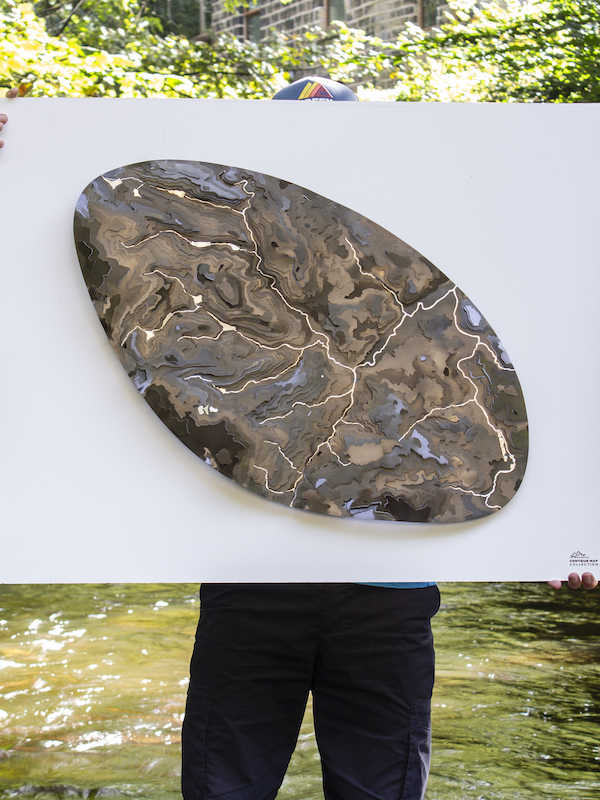 Photograph of someone hidden behind a white square that they are holding. You can only see the top of a cap in the centre their feet at the bottom on finger tips on the sides. In the centre of the white square is a large pebble shape of greys, browns and blues that shows a contour map of a river basin.