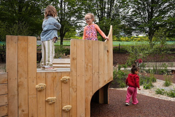 Photograph of a children's outdoor play area. Two young children stand on top of a wooden structure in the background a young child walks toward trees and bushes and a low level planing area. 