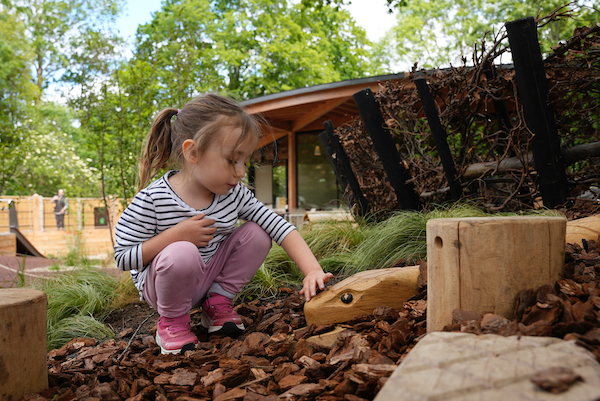 Photograph of a young girl crouching down in an outdoor play area. She wears pink trousers and top, her left hand is outstretched as she pets a wooden snakes head laying on the ground amongst the wood chip.