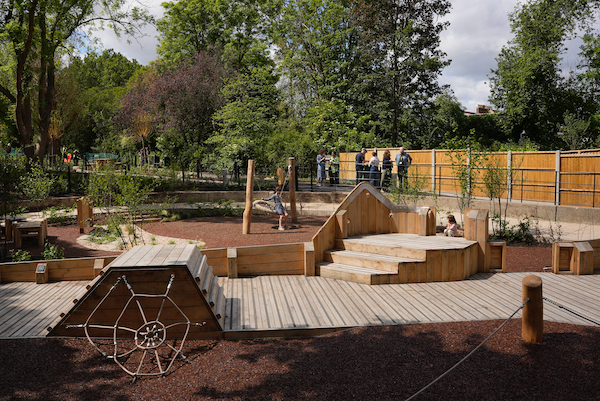 Photograph of an outdoor play area, a climbing roped area is in the shape of a spiders web. There are wooden walkways and play spaces set amongst trees and green bushes. 