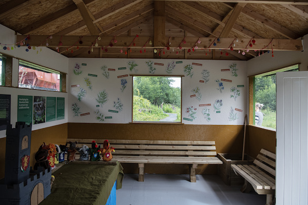 Photograph of the inside of an outdoor classroom. There are wooden benches and windows that look out on to green bushes. On the walls are illustrations and names of plants.  