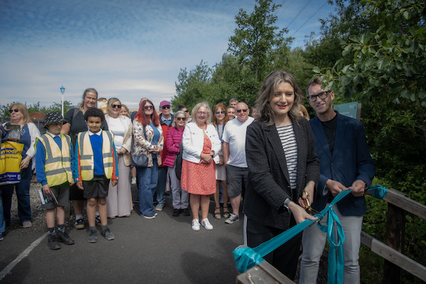 A photograph of a man and a woman cutting a red ribbon outside. Behind the two people stand member so the public and school children they are smiling. There is blue sky and green bushes behind the crowd. 