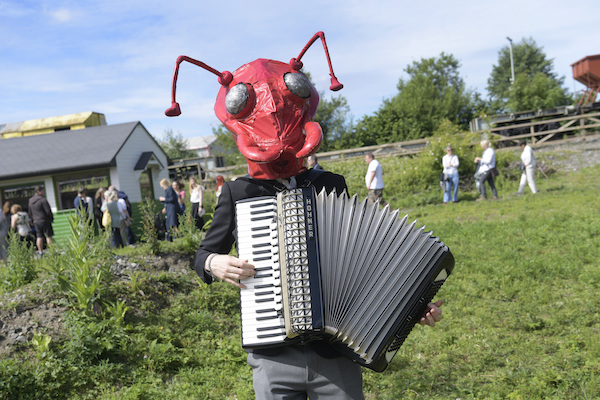 Photograph of a musician looking at the camera, he is playing an accordion and he wears a mask of a red insect which covers his face. In the back ground visitors walk in the woodland walk. 