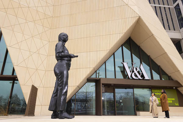 Photograph outside the entrance to V&A East a large 18ft bronze sculpture of a woman dominates the frame to the right stand two women looking up at the sculpture. 