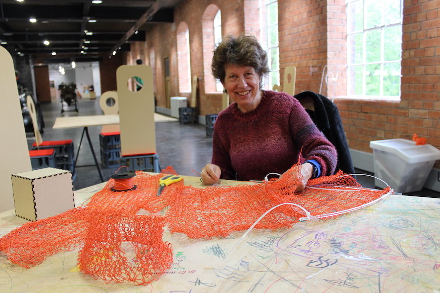 A woman sits at a table in a large open room, on the table in front of her is some orange netting and scissors the woman is smiling.