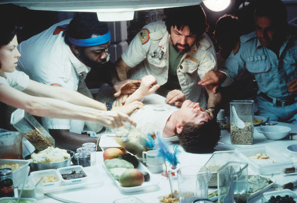 Film still from Alien. A man lays on a table surrounded by bowls and food. Four other people surround him with concern. They wear uniformed shirts. There are three men and one woman reaching out to him.