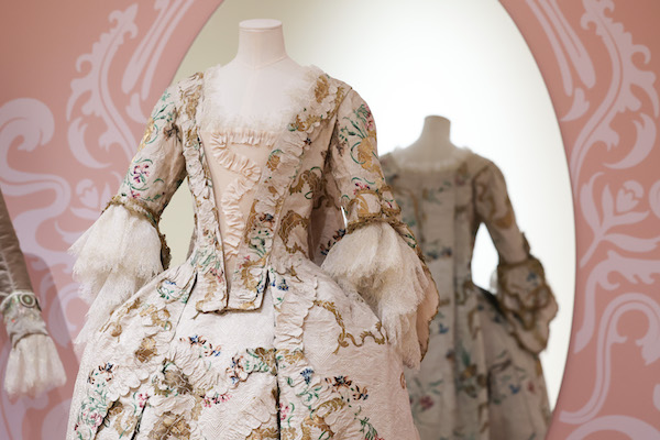 Photograph of an 18th century dress with bodice and skirts. It features lace on the sleeves and round the neckline. It is a cream colour with ruffles and flower patterns. 