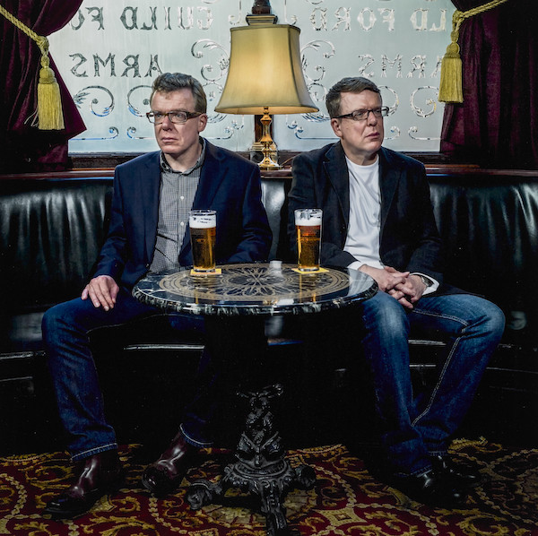 Two middle aged men sit in a pub at a small round table, in front of them on the table both have a pint of beer. The men are twins they are brothers from the band The Proclaimers. They have short hair and both wear glasses, suit jackets and blue jeans with open necked shirts.. They are looking off to the left and right of the camera. The pub floor is heavily patterned, there is a lamp behind them and etched glass.