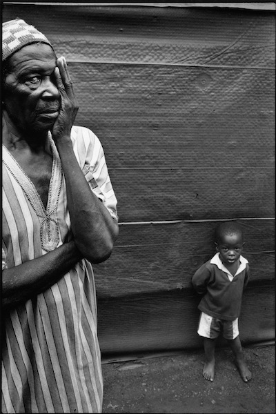 A black and white portrait photograph of an older black man one arm is crossed in front of his body his other arm is on his face covering his left eye. He looks directly at the camera with his uncovered eye. He wears a striped tunic and cap style hat. Behind him stands a small black child with bare feet who also looks directly at the camera. He wears shorts and a top he stands up against what looks like a temporary structure. 