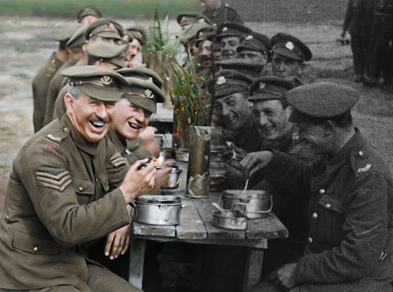 A photograph shows a long wooden table men in uniform sit either side eating, drinking and laughing. They are outside in the background there is grass. They eat from round silver tins. The right hand side of the photo is in black and white. The left hand side is in colour. You can see the uniforms are green.