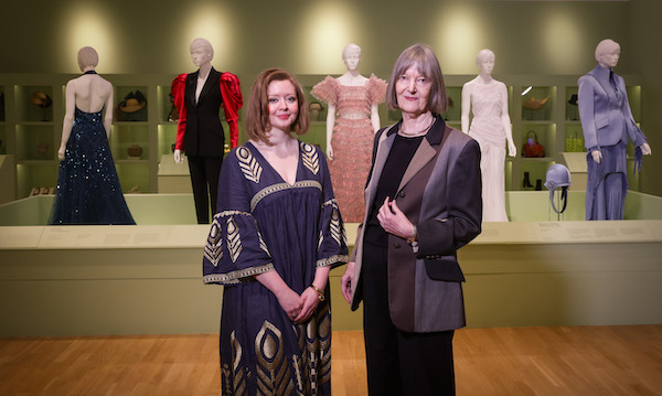 Photograph of two women standing in front of a display of clothing in a museum. The woman on the left is smiling and wearing a dark blue dress with gold patterns. The older woman on the right wears black trousers and top with a brown/beige suit jacket.
