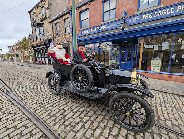 On a cobbled street in front of traditional shops a black vintage car is parked. At the back sits Father Christmas in a red and white suit with a big white beard and christmas hat. He is driven by a man wearing a dark suit and red scarf.