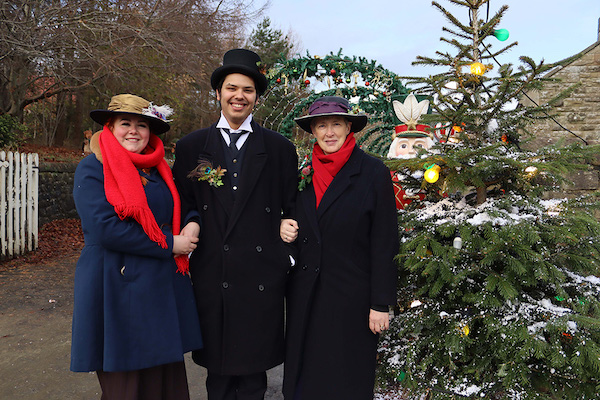 Three people, two women and one man stand outside next to a fir tree sprinkled in white snow. The link arms and smile at the camera, they wear warm winter clothes and hats. Their clothes are period dress from the 1820s to the 1950s.