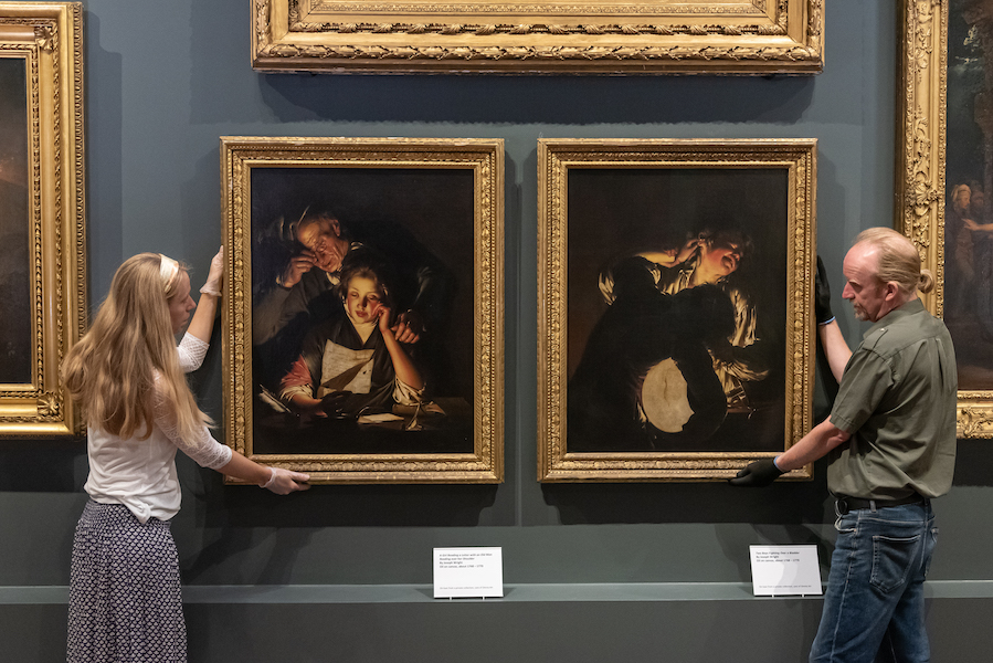 Two large oil paintings with gold frames hang on a gallery either side of the paintings stand a female and male curator their hold their hands up to the frames.