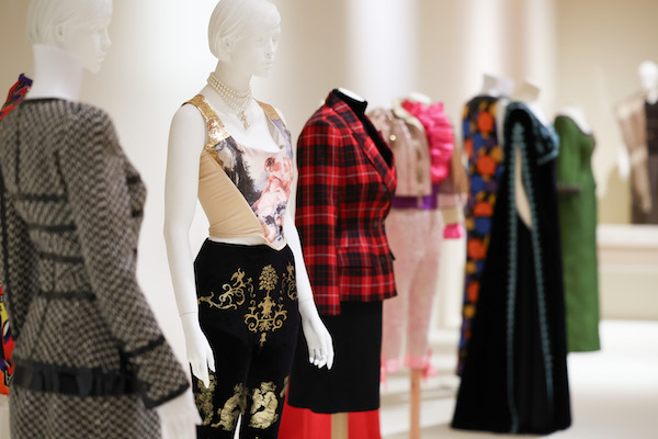 Photograph of a costume display in a museum. A number of outfits are on display slightly out of focus. In focus is a outfit with black and gold trousers with a cream bodice. The mannequin has pearls around its neck. 