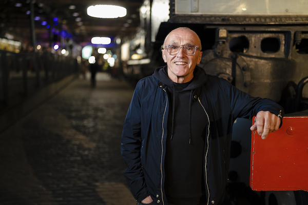 A white older man with clear glasses and a bald head smiles at the camera. He wears a blue jacket and his arm rests on a train. Behind him in the distance is a museum exhibition space with trains. 