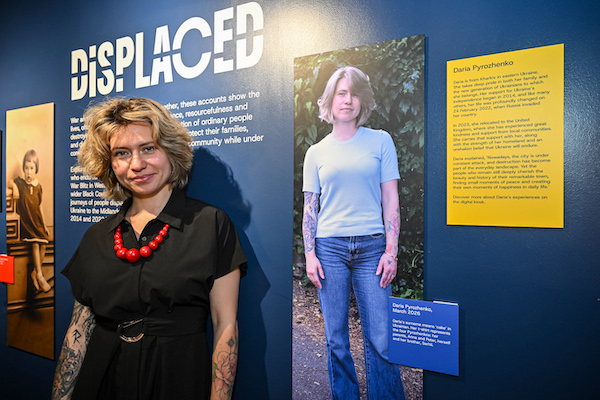 Photograph of a young woman with blonde medium length hair, wearing a black shirt and red beads. She is standing beside a photograph of herself in an exhibition display. 