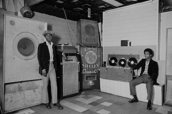 Black and white photograph of two black men in the corner of a room edged with amplifiers and speakers. One speaker seen in the centre of the image is labelled 'COUNT SHELLY Sounds'. On the right hand side of the image, one of the men is sat on a bench looking directly at the camera, his right arm resting on a large box of records. The other man stands next to a very large speaker wearing flared trousers and a cloth cap.