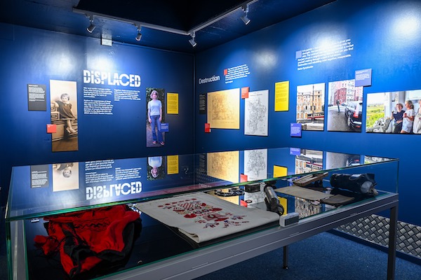 Photograph of an exhibition, there is a glass topped display table with objects in the foreground and blue walls feature text and photographs