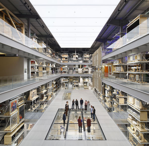 Photograph of the inside of a museum store, the central hall is open and has a square section of see-through floor in the middle around which stand a crowd of people. The shelves of the store are full of museum objects. 