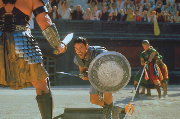 Film still from Gladiator. Two Gladiators fight in an arena, we see one man partially from behind, he wears armour and carries a sword in his right hand. In front is another man, mouth open ready to charge forward, he has a sword in his right hand and round shield in his left. 
