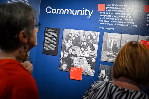 Photograph of the back of two women as they read wall text and look at photographs in an exhibitions. 