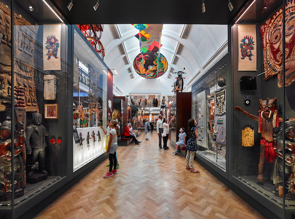 Photograph of a museum gallery. Brightly colour exhibits in museum display cases frame a walkway with a wooden floor. Visitors stand along the walkway looking at the exhibits. 