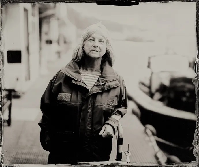 A black and white photo of an older woman, she stares straight at the camera. She has light coloured hair cut into a shoulder length bob. The background is blurry but it looks to be by a quayside with a boat behind her on her left. There is a metal railing on the side of the quay that she slightly leans on.