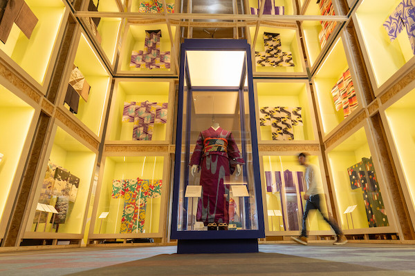 Photograph of the inside of the exhibition, there are floor to ceiling glass display cabinets with yellow walls, the kimonos are displayed in them they are all different colours their arms outstretched. In the centre of the image is a free standing glass display showing a red kimono worn on a mannequin.